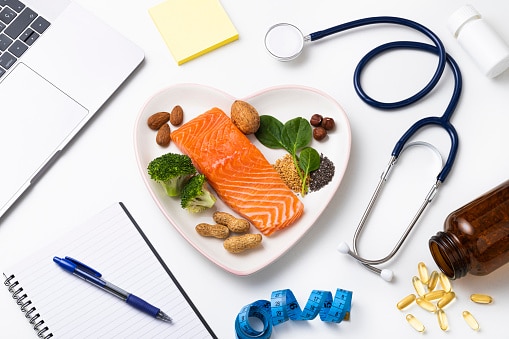Top view of a heart shaped plated full of food rich in omega-3 such as a salmon fillet,
spinach, chia and flax seeds, broccoli, almond, peanut and hazelnuts. The plate is at the center of the image and it is surrounded by a stethoscope, a tape mesure, a notepad and some omega-3 capsules.
