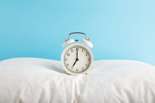 White alarm clock on pillow at light blue wall background. Pastel color. 7 o'clock in morning. Waking time concept. Closeup. Front view.