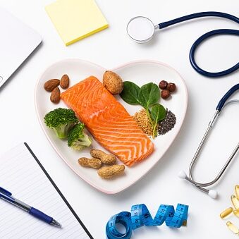Top view of a heart shaped plated full of food rich in omega-3 such as a salmon fillet,
spinach, chia and flax seeds, broccoli, almond, peanut and hazelnuts. The plate is at the center of the image and it is surrounded by a stethoscope, a tape mesure, a notepad and some omega-3 capsules.
