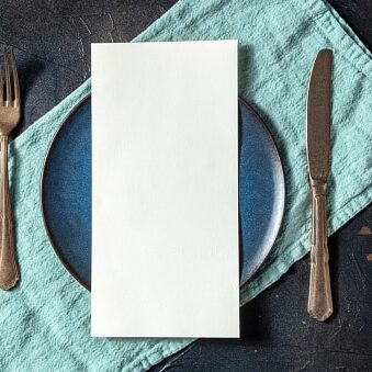 Dinner menu or invitation concept. A piece of white paper on a set table, with a fork, a knife, and a blue plate, overhead flat lay shot on black