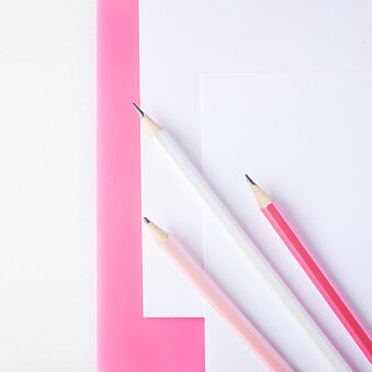 Three pencils lie on a pink clipboard and a sheet of paper. in the frame you can see a plant in a cache-pot